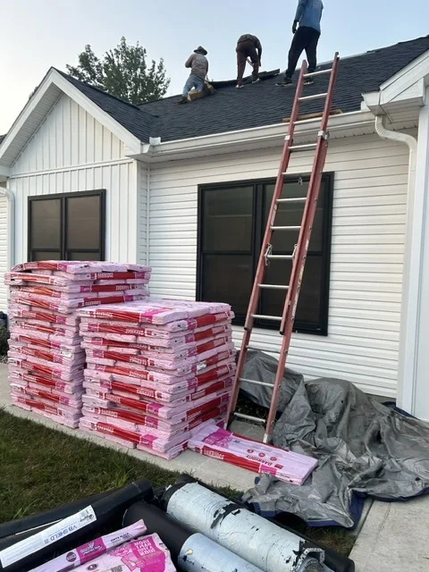Roofing crew on a white farmhouse roof with shingle bundles and underlayment staged below
