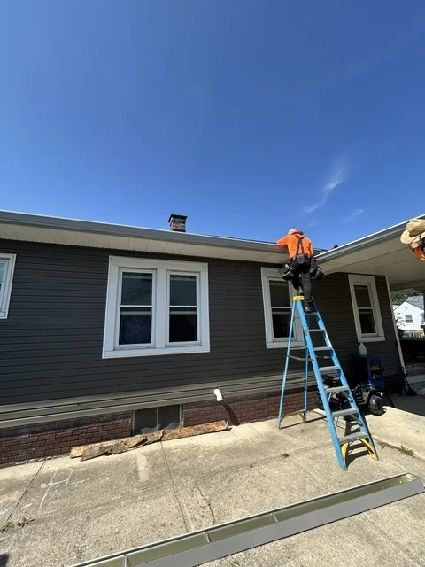 Crew member on a ladder working at the roof edge of a gray-sided home
