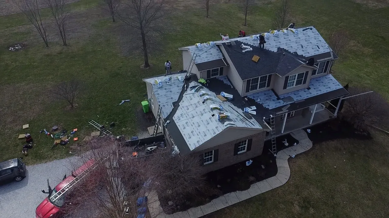 Drone view of a large two-story home during a roof replacement, with much of the roof in white synthetic underlayment, the crew on top and a dump trailer and trucks in the driveway