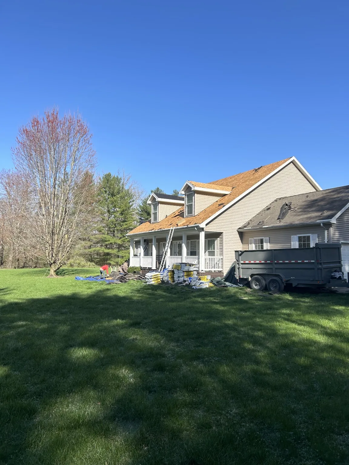 The same dormered Cape Cod home mid-replacement, the main roof stripped down to clean OSB decking with a dump trailer and materials staged in the yard