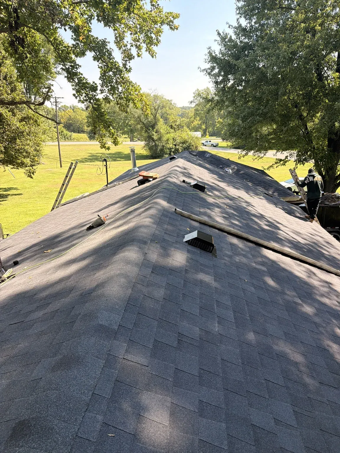 Looking up a shingle roof with box vents on a sunny day, with a roofer working near the far end of the ridge