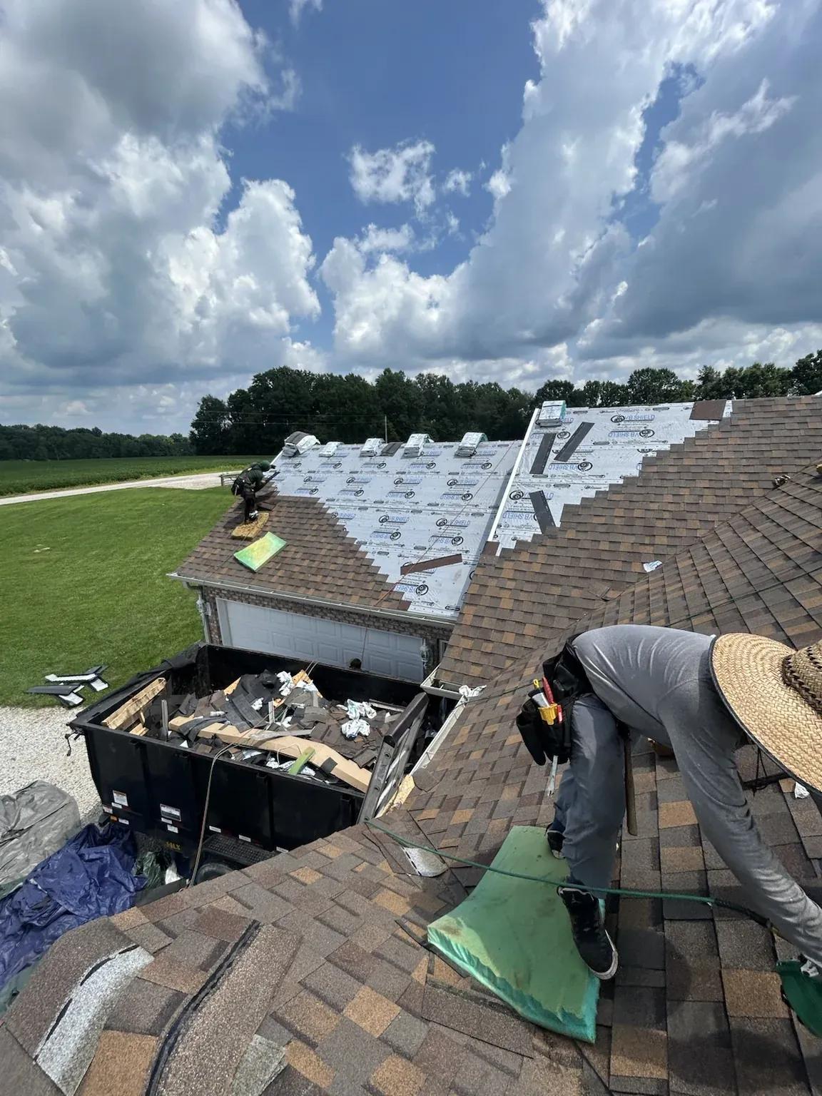 Roofer working section by section beside a dormer, with the upper section already in white underlayment and a dump trailer of tear-off debris below