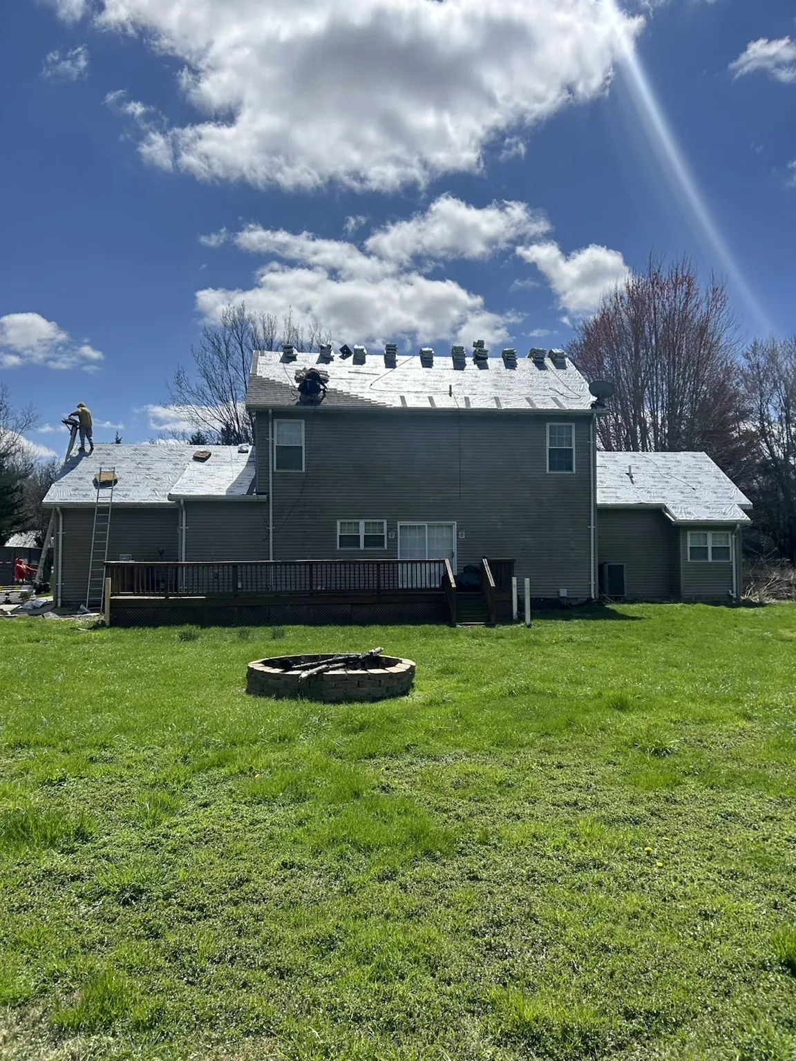 Back of a two-story home with a deck and fire pit, the roof covered in white synthetic underlayment with shingle bundles staged along the ridge