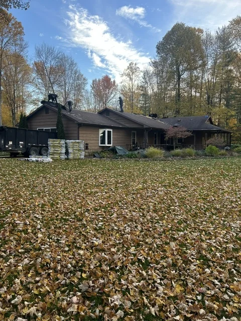 Dark-sided home in the fall with shingle bundles and a trailer staged for a roof replacement